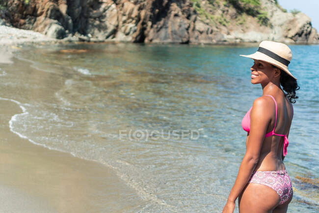 Side view of cheerful ethnic female tourist in swimwear and hat looking away on ocean coast — Stock Photo