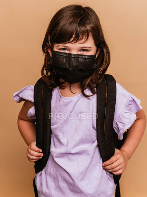 Приємна дитина з рюкзаком і захисною маскою від коронавірусу на коричневому тлі в студії — стокове фото