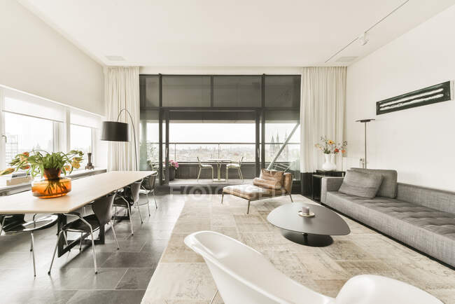Interior de la moderna y espaciosa sala de estar con cómodo sofá y mesa de madera y sillas en un lujoso apartamento contemporáneo con ventanas panorámicas - foto de stock