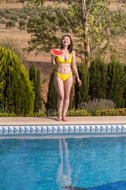 Vista lateral de una alegre hembra étnica en bikini amarillo parada cerca de la piscina y comiendo sandía fresca en un día soleado en verano mientras mira la cámara - foto de stock