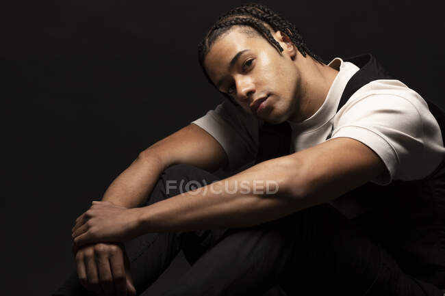 Side view of confident young ethnic hipster male model with braided hair wearing white shirt with black clothes looking at camera while sitting in dark studio — Stock Photo