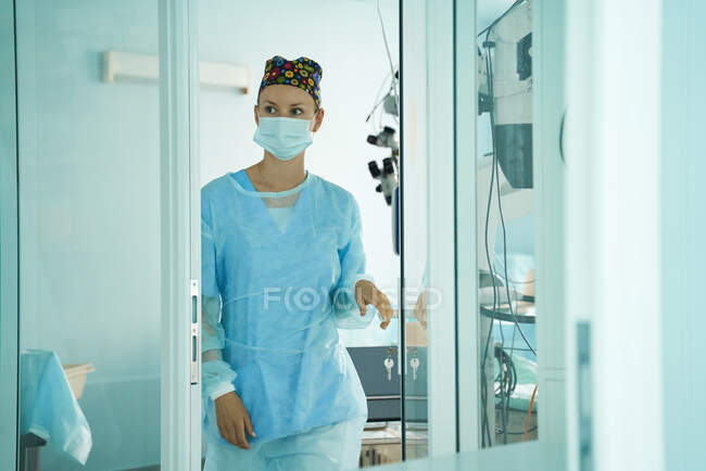 Alegre médico adulto femenino en máscara estéril y gorra ornamental mirando hacia otro lado en el hospital - foto de stock