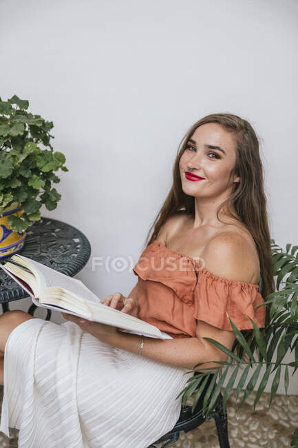 Side view of graceful female in skirt leaning legs on chair while chilling in patio in summer and enjoying interesting story in book at weekend — Stock Photo