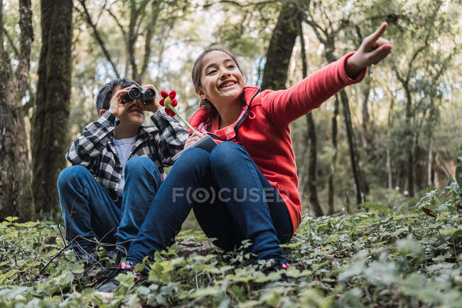 Menina étnica escrevendo em bloco de notas contra irmão olhando através de binóculos enquanto sentado em terra em florestas de verão — Fotografia de Stock