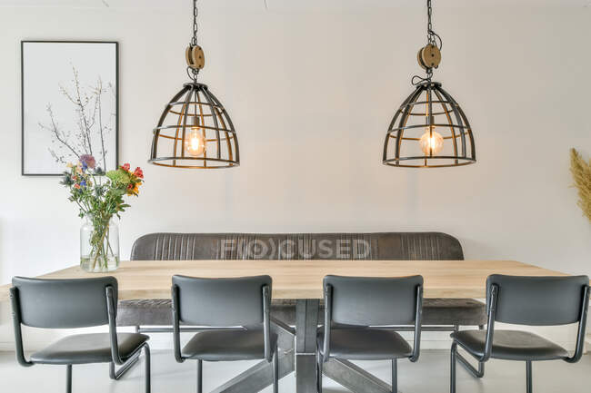 Элегантный интерьер современной столовой со стульями и диваном рядом с деревянным столом с букетом цветов под светящимися лампами с металлическими элементами в квартире в стиле лофт — стоковое фото