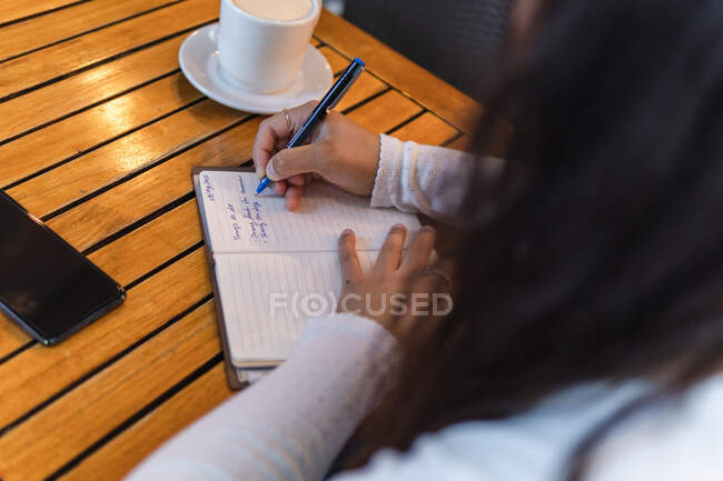 Cropped unrecognizable ethnic businesswoman in casual clothes taking notes in planner while sitting at table with cup of coffee and smartphone in cozy cafeteria with green plants — Stock Photo