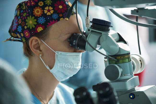 Доросла жінка-лікар у стерильній масці та декоративній медичній кепці, яка дивиться через хірургічний мікроскоп проти колеги по сільськогосподарській галузі в лікарні — стокове фото