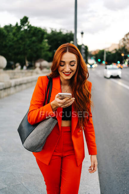 Mujer sonriente con el pelo rojo y en traje naranja grabando mensaje de audio en el teléfono móvil mientras se comunica con un amigo en las redes sociales y caminando en la calle de la ciudad - foto de stock