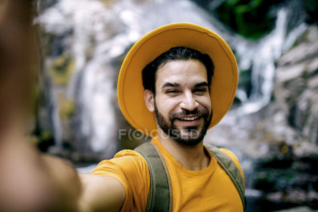 Viajante do sexo masculino encantado em roupa amarela tirando selfie no fundo enterrado de rochas durante o trekking na floresta — Fotografia de Stock