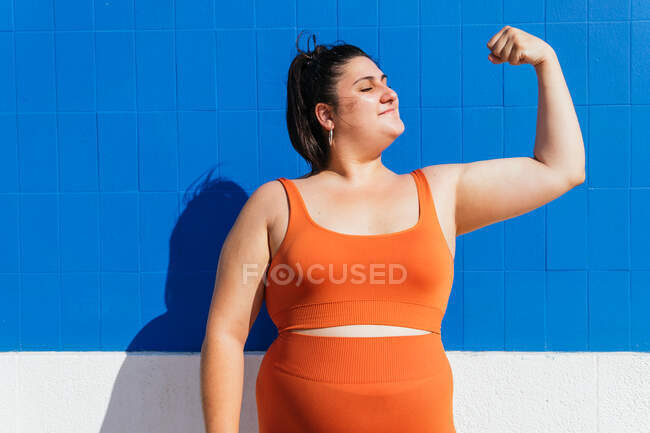 Potente atleta femenina de talla grande en ropa deportiva demostrando músculos mientras mira hacia otro lado contra la pared de azulejos azules en la calle - foto de stock