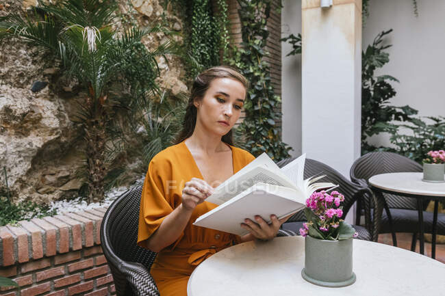 Heitere zarte Frau liest interessantes Buch, während sie am Tisch auf der Sommerterrasse des Hauses sitzt — Stockfoto