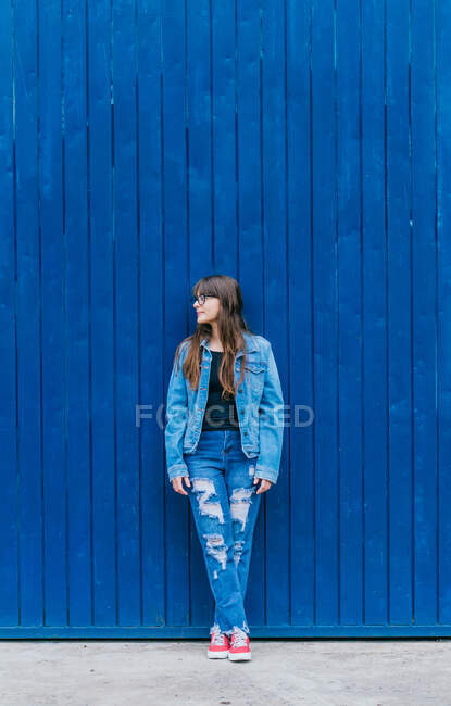 Модная хипстерша в джинсовой куртке и джинсах, опирающаяся на голубую стену на городской улице и отворачивающаяся — стоковое фото