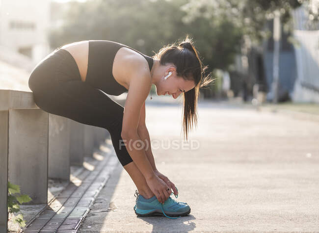 Vista lateral de atleta mujer atando cordones en zapatillas y mirando hacia abajo mientras se prepara para el entrenamiento de fitness en la calle - foto de stock