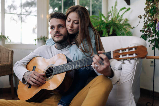 Alegre tatuado masculino músico tocando guitarra perto de conteúdo fêmea amado enquanto olhando para o outro em poltrona em casa quarto — Fotografia de Stock
