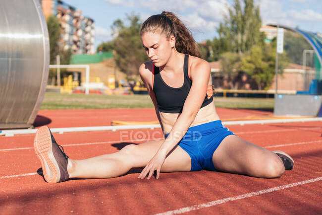 Концентрированная спортсменка, сидящая на стадионе и растягивающая ноги во время легкой атлетики — стоковое фото