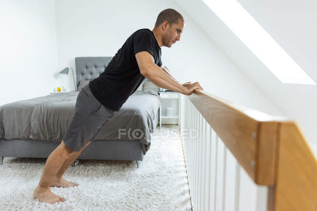 Seitenansicht eines Mannes, der Liegestütze in der Nähe von Holzgeländern macht, während er morgens im Schlafzimmer trainiert — Stockfoto