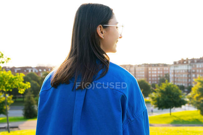 Rückansicht einer Frau in trendiger Kleidung, die an einem sonnigen Tag auf einem grünen Hügel vor dem Hintergrund der Stadt steht und wegschaut — Stockfoto