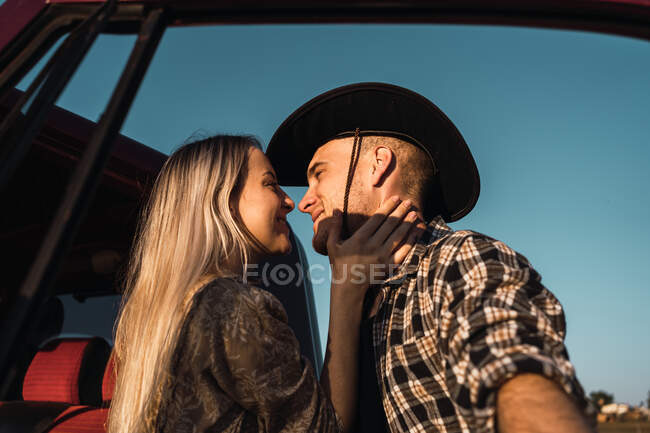De baixo vista lateral da jovem mulher amorosa beijando o homem em chapéu de cowboy ternamente perto do carro no fundo do céu azul à noite — Fotografia de Stock