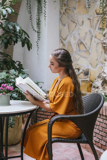 Seitenansicht der heiteren zarten Frau liest interessantes Buch, während sie am Tisch auf der Sommerterrasse des Hauses sitzt — Stockfoto