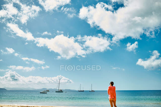 Visão traseira de turista feminino envolto em roupa rosa enquanto passa o tempo na praia de areia contra o mar azul-turquesa com iates e céu azul nublado em Fuerteventura, Espanha — Fotografia de Stock