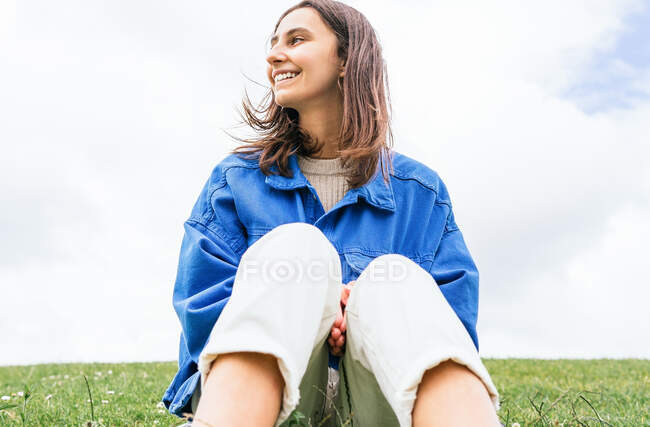 Low angle of glad female sitting on hill in nature on windy day while looking away — Stock Photo