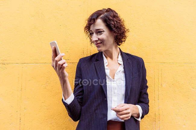 Auto asegurado elegante mujer usando traje formal de pie mirando a la pantalla del teléfono móvil - foto de stock