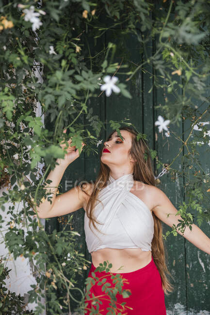Mulher graciosa com lábios vermelhos e em roupa de verão desfrutando de aroma de flores aromáticas crescendo no pátio da casa — Fotografia de Stock