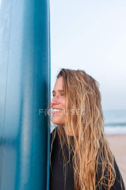 Vista lateral de surfista femenina feliz de pie con tabla SUP azul en la orilla del mar de arena en verano y mirando hacia otro lado - foto de stock