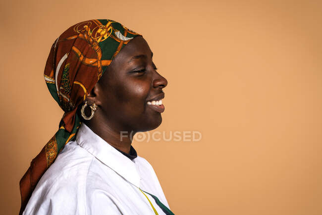 Веселая афро-американская модель в модном платке и белой рубашке смеялась с закрытыми глазами на бежевом фоне в студии — стоковое фото