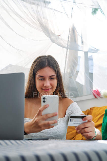 Jeune femme de contenu assis sur la table et effectuant le paiement avec une carte en plastique pour commander lors des achats en ligne sur téléphone mobile — Photo de stock