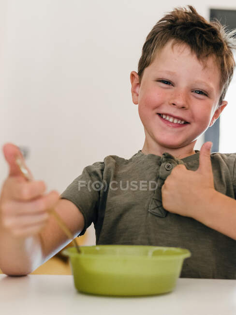 Entzückter Junge isst leckeres Essen, während er am Tisch in der Küche sitzt und wie eine Geste zeigt — Stockfoto