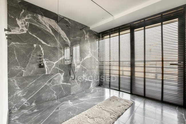 Інтер'єр сучасної просторої ванної кімнати з душовою одиницею на плитці з мармуровою стіною зі скляною перегородкою біля затіненого вікна в сучасній розкішній квартирі — стокове фото