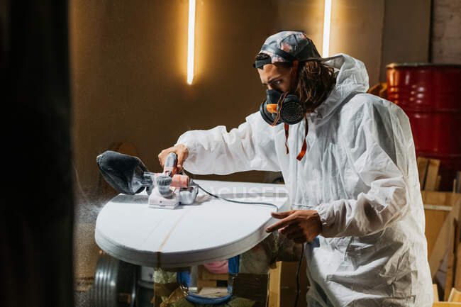 Male shaper in protective respirator and costume using electric planer and polishing surface of surfboard in workshop — Stock Photo