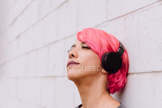 Young female with bright pink hair listening to music with headphones while standing near white wall with eyes closed — Stock Photo