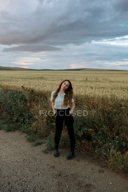Молода усвідомлена жінка з закритими очима на дорозі біля лугу під хмарним небом увечері в сільській місцевості — стокове фото