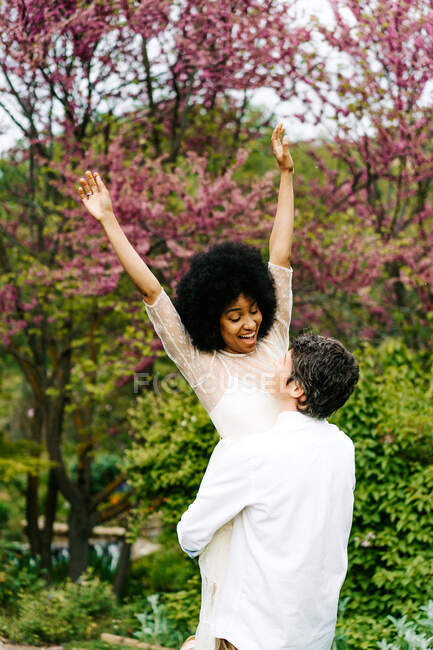 Vista lateral del hombre levantando mujer negra mientras está de pie en el prado en el jardín - foto de stock