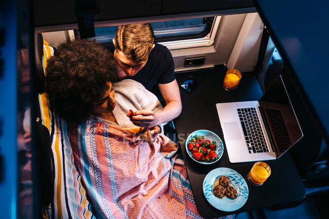 From above of happy multiracial couple having meal near laptop as resting together in camper van during vacation — Stock Photo