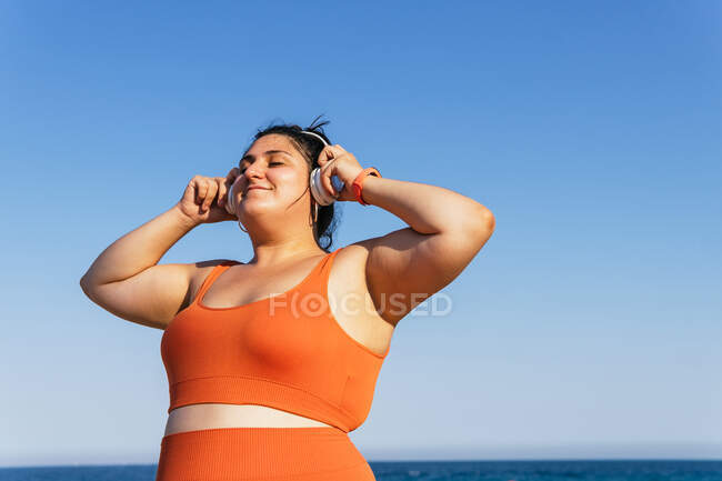 Весела етнічна жінка-спортсменка з пишним тілом слухає пісню з навушників, закриваючи очі під блакитним небом — стокове фото