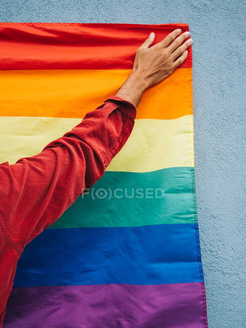 Cropped anonymous homosexual male standing with LGBT rainbow flag near blue wall in city street — Stock Photo