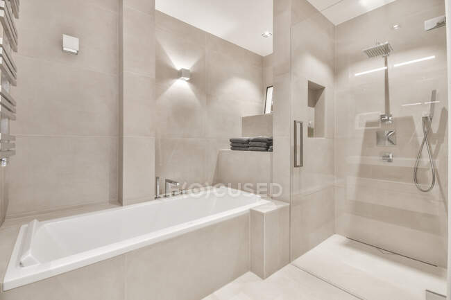 Современный дизайн интерьера светлой ванной комнаты с белой ванной и просторной душевой кабиной, украшенной серой плиткой с подсветкой — стоковое фото