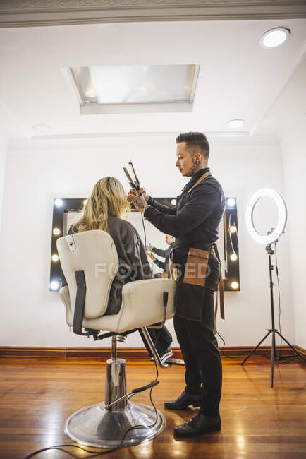 Cabeleireiro masculino usando ferro para enrolar fechaduras loiras de cliente feminino durante o trabalho no salão de beleza — Fotografia de Stock