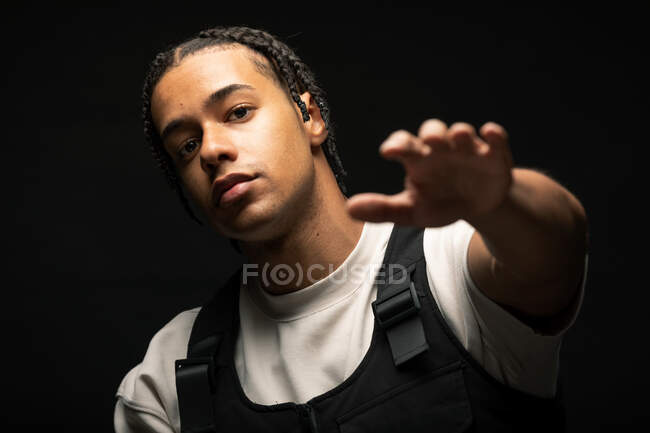 Guapo joven étnico masculino con trenzas afro vestido con ropa blanca y negra mirando a la cámara mientras está sentado en un estudio oscuro - foto de stock