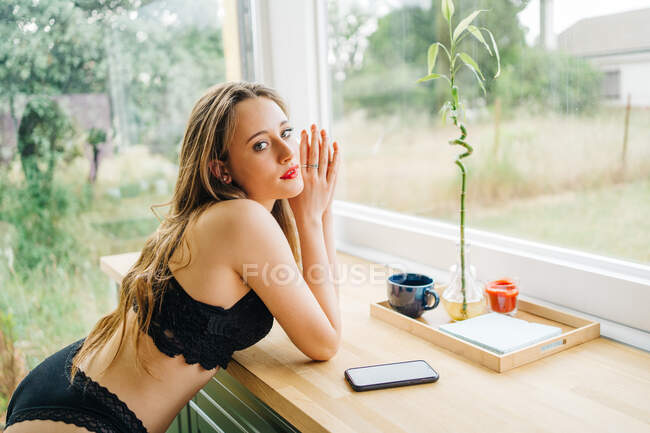 Vista lateral de seductora hembra en lencería negra y con labios rojos sentada en mostrador en cocina en casa y mirando cámara - foto de stock
