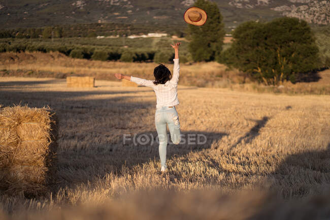 Back view of unrecognizable female throwing hat and running along dried field in summer evening in countryside — Stock Photo