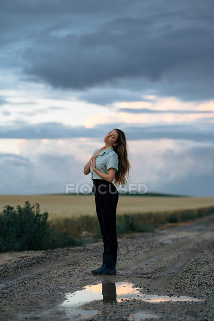 Стильна молода жінка торкається довгого волосся на проїжджій частині, відбиваючись в калюжі під хмарним небом в сутінках — стокове фото