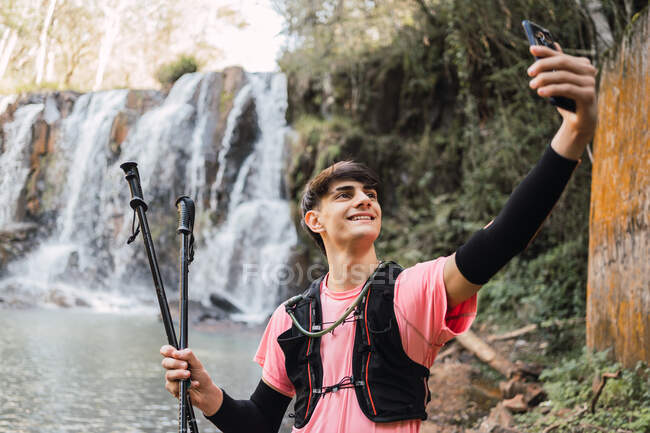Senderista masculino sonriente tomando uno mismo tiro en el teléfono inteligente mientras está de pie en el fondo de la cascada y el lago en los bosques durante el trekking - foto de stock