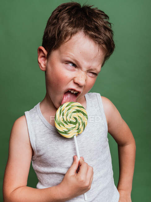 Preteen menino sorrindo e lambendo saboroso pirulito espiral no fundo verde no estúdio enquanto olha para longe — Fotografia de Stock
