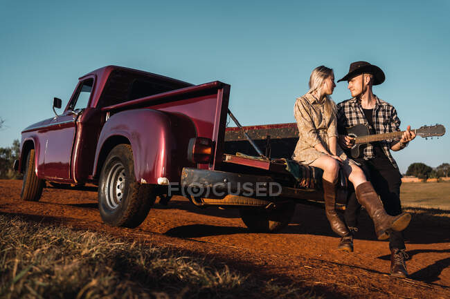 Парень в ковбойской шляпе играет на акустической гитаре, сидя с девушкой в багажнике красного пикапа, припаркованного на песчаной дороге в сельской местности. — стоковое фото