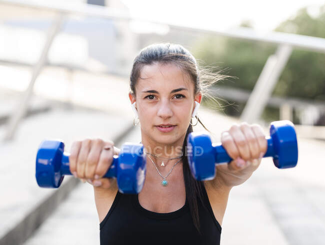 Determined female athlete doing exercise with dumbbells during fitness workout on city street in summer — Stock Photo