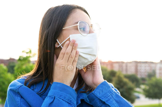 Female in glasses putting on protective medical mask during coronavirus outbreak in city and looking away — Stock Photo